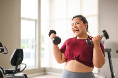 Genç, pozitif, gülümseyen, neşeli, kilolu kadın spor salonunda dambıllarıyla antrenman yapıyor. Spor kıyafetli şişman kadın spor yapıyor. Kilo verme egzersizi, sağlıklı yaşam tarzı konsepti