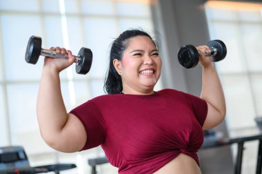 Genç, pozitif, gülümseyen, neşeli, kilolu kadın spor salonunda dambıllarıyla antrenman yapıyor. Spor kıyafetli şişman kadın spor yapıyor. Kilo verme egzersizi, sağlıklı yaşam tarzı konsepti