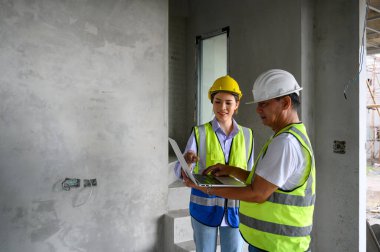 Yelekli, kasklı, dizüstü bilgisayarlı genç ve çekici bir inşaat işçisi. İnşaat alanında duruyor. Ev yapımı projesi. Mühendis ustabaşı iş yerinden bir iş arkadaşıyla konuşuyor.