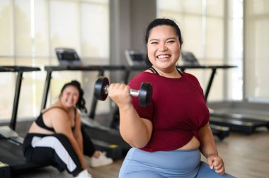 Genç, pozitif, gülümseyen, neşeli, kilolu kadın spor salonunda dambıllarıyla antrenman yapıyor. Spor kıyafetli şişman kadın spor yapıyor. Kilo verme egzersizi, sağlıklı yaşam tarzı konsepti