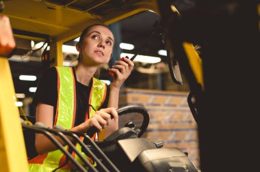 Depo, radyo ve nakliye deposunda forklift. Güvenlik üniformalı kadın nakliye işçisi telsizle konuşuyor, yük deposunda forklift kullanıyor.