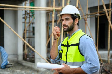 Beyaz miğferli çekici bir inşaat işçisi inşaat alanında dururken telsiz kullanıyor ve plan tutuyor. Ev yapımı projesi