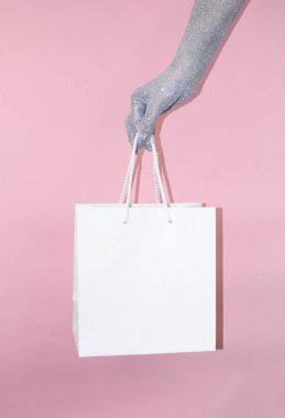 Işıl ışıl bir el, parlak pembe bir arka fonun önünde sade beyaz bir alışveriş torbasını kavrıyor..