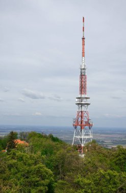 Uzun, kırmızı ve beyaz radyo kulesi yeşil ağaçlarla kaplı tepede duruyor..