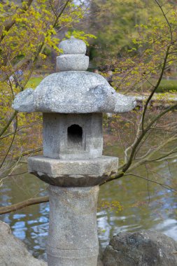 Etrafı yeşilliklerle çevrili, dingin bir göletin yanında duran bir taş fener..