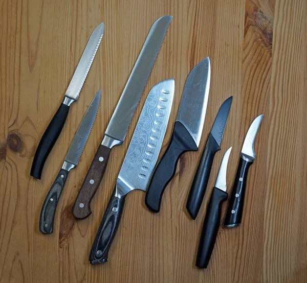 Eight kitchen knives on wooden board. The first is a tomato knife, the third a bread knife. Next a Japanese Gyuto knife.