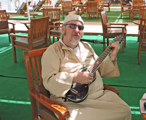 A European man sits on a chair on the deck of a Nile cruise ship and plays a black travel guitar. He is dressed in Egyptian style.
