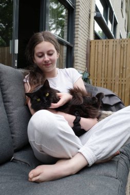 14 yaşındaki bir kız siyah rakun kedisine sarılıyor. Kanepede bacak bacak üstüne atmış oturuyor.