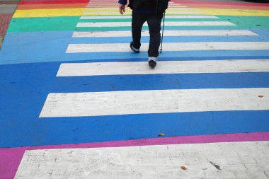 Bastonlu bir adam karşıya geçiyor. Zebra geçişi gökkuşağı renkleriyle boyanmış.