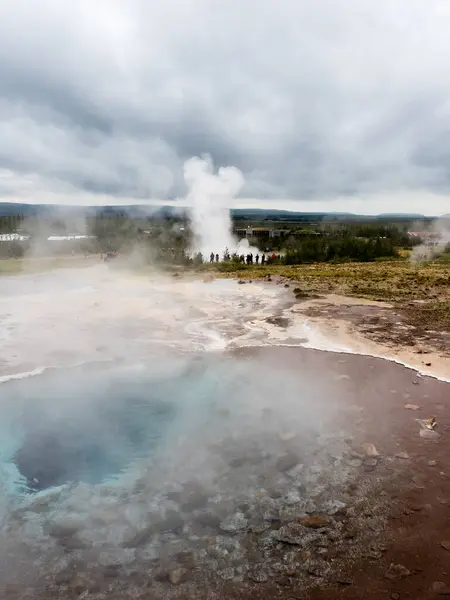 Steaming colorful hot spring pool with Strokkur geyser at the background in Geysir geothermal area, Iceland