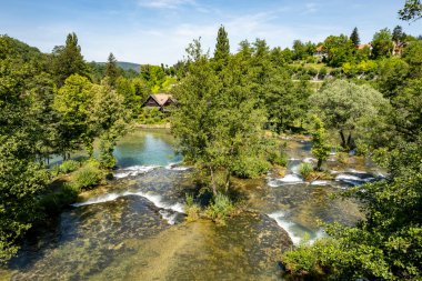 Rastoke şelalelerindeki Korana nehrinin güzel çağlayanları ve nehir kenarındaki ahşap kulübeler, ağaçlar ve bitkilerle kaplı.