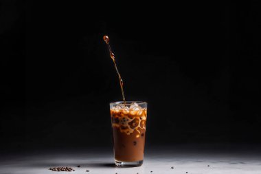 Bir buzlu kahve bardağıyla koyu bir arka plan. Buzlu camdan görülen zengin kahverengi kahve görsel olarak çarpıcı bir kompozisyon oluşturur. Kahvenin içinde buz parçaları yüzer, ferahlatıcı bir dokunuş katar. Karanlık, neredeyse siyah arkaplan Visua 'yı güçlendirir.