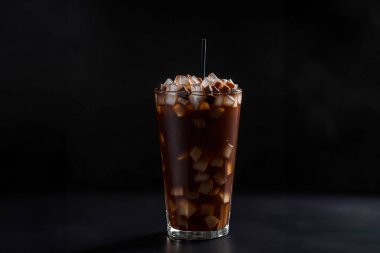 Bir buzlu kahve bardağıyla koyu bir arka plan. Buzlu camdan görülen zengin kahverengi kahve görsel olarak çarpıcı bir kompozisyon oluşturur. Kahvenin içinde buz parçaları yüzer, ferahlatıcı bir dokunuş katar. Karanlık, neredeyse siyah arkaplan Visua 'yı güçlendirir.