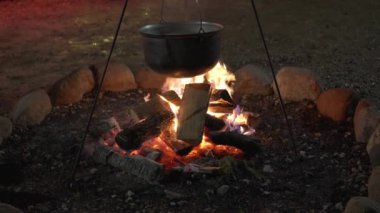 Geceleri üzerinde bir tencere asılı duran şenlik ateşi, yemek pişirme, arka planda ışıkları olan dekore edilmiş bir Noel ağacı, tripodsuz çekilmiş bir sahne, bir ev videosu hissi yaratıyor.