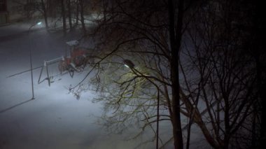 Kar şiddetli yağıyor, gece kar yağıyor, fener ışıkları ile aydınlatılıyor, iki lamba, yapraksız ağaçlar ve bir çocuk parkı, bir apartman dairesi, el kamerasıyla çekilmiş