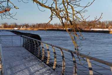 Üzerinde dallar asılı bir köprünün kış manzarası, diğer kıyılar karla kaplı, su donmamış, Jelgava Kalesi yakınlarında nehir görülebilir.