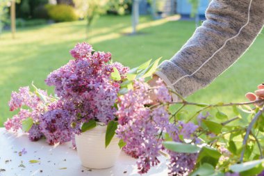 Bahçedeki beyaz bir masadaki vazoda leylak çiçeği aranjmanı, ışığa yakın mesafeden çekilmiş farklı odak, bulanık hareket, gün batımı ışığı.