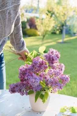 Bahçedeki beyaz bir masadaki vazoda leylak çiçeği aranjmanı, ışığa yakın mesafeden çekilmiş farklı odak, bulanık hareket, gün batımı ışığı.