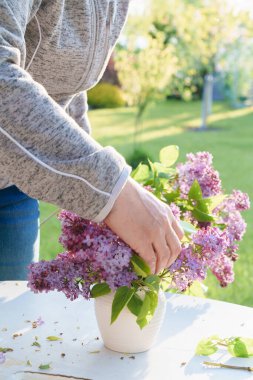 Bahçedeki beyaz bir masadaki vazoda leylak çiçeği aranjmanı, ışığa yakın mesafeden çekilmiş farklı odak, bulanık hareket, gün batımı ışığı.