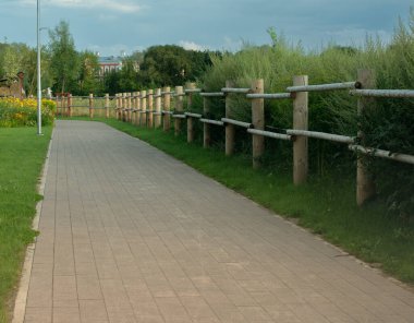 Parkın içinde kıvrılan kaldırımlı bir yolun basit ve manzaralı bir görüntüsü. Yol kenarlarda doğal ahşap bir çitle çevrili, arkasında uzun otlar ve diğer bitkiler yetişiyor. Bu bir huzur ve rahatlama hissi yaratır. Doğa temalı u için ideal