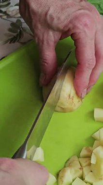Bir insanın elinin, yeşil bir kesme tahtasının üzerinde patatesi küçük, üniformalı küplere ayırmak için bıçak kullanması.