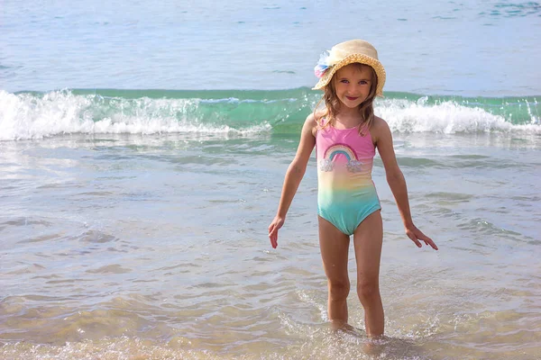 在一个阳光灿烂的日子里 一个身穿鲜艳泳衣 头戴一顶帽子的漂亮小女孩站在大海的背景下 专心致志地注视着大海 — 图库照片