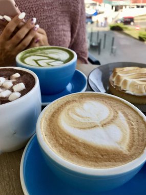 Kahve bardakları cappuccino, latte ve kakao, pastalar ve kafenin arka planında telefonu olan bir kızın elleri.