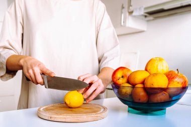 Kadının elleri taze limon içeceği yapmak için tahta keserek limon dilimleri kesiyor. Bir kâse taze meyve yakınlarda duruyor.