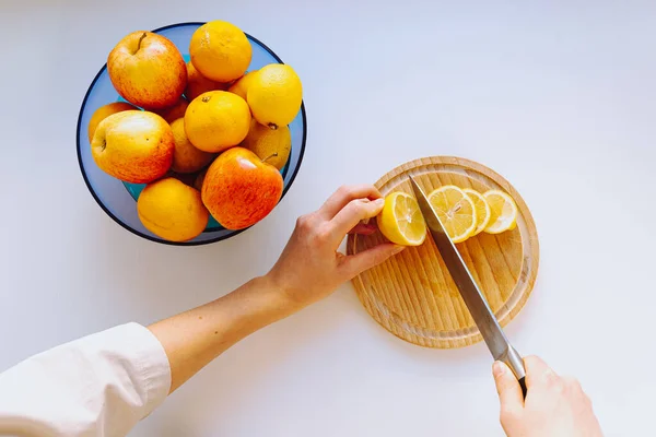 woman\'s hands cut lemon slices on cutting wooden board to make refreshing lemon drink, bowl of fresh fruits stands nearby, top view. preparation of fruit slices, vitamin fruit salad