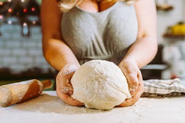 Bukleli kadın, ev hanımı, elinde un serpiştirilmiş hamuru mutfak masasında, Noel süsleriyle süslenmiş mutfağın bulanık arka planında tutuyor. Tatil hamuru pişirmek, pizza, lezzetli turta, evde, ev mutfağında.