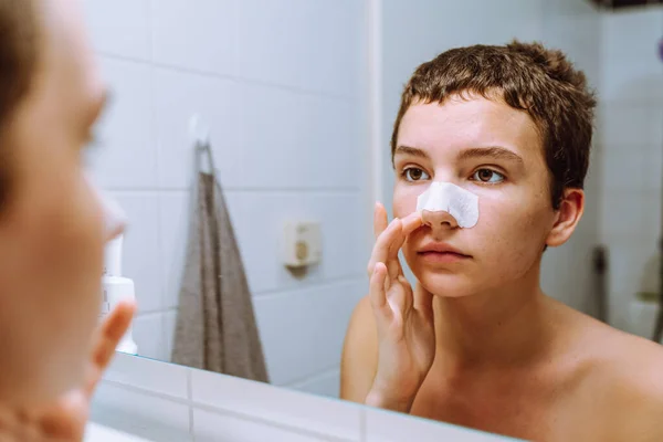 teenage girl, with short haircut, looks at reflection in mirror, on nose there is cosmetic patch for acne and black dots. teenage cosmetics, facial skin care, self-recognition