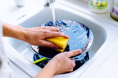 Kadınların elleri musluk suyunun altında bulaşık süngeriyle kirli tabakları yıkıyor. Yemekten sonra bulaşık yıkama konsepti, ev işleri, mutfak lavabosu, aşırı su tüketimi, bulaşık makinesi arızası. Ev hanımı mutfak lavabosunda bulaşık yıkıyor.