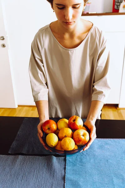 Attractive young woman with  short haircut, in smart brown dress, puts bowl of fruit on table. Home life, teenager girl at home in dining room puts bowl of citrus fruits in middle of table