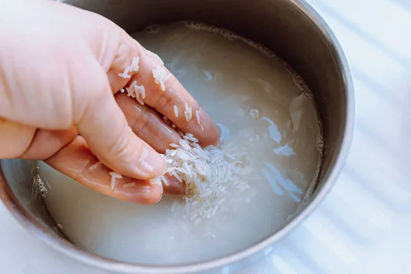 rinsing rice before cooking. hand of cook washes rice grains in metal ladle under running water. technology of recipe for correct preparation of rice step by step