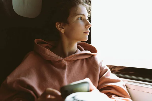 teenage girl in warm pink sweatshirt sits near window in train car, looks out window. Place to test, journey alone. Toned photo. Student teenager girl travels alone, nostalgia, sadness loneliness, loss, depression