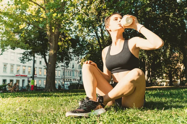 teenager girl of athletic muscular build sits on lawn in city park, after sports workout, yoga, drinks tonic energy drink isotonic for athletes. Workout stretching sports on coast in summer in park