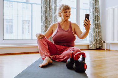 Orta yaşlı sarışın kadın, iri yapılı, iri göğüslü, spor tişörtlü, yoga yapıyor, selfie çekiyor, mindere oturuyor, parke zeminli parlak güneşli bir odada, geniş pencerenin bulanık arka planına karşı. Yoga vücut pozitifliği evde yetişkin bir kadın için.