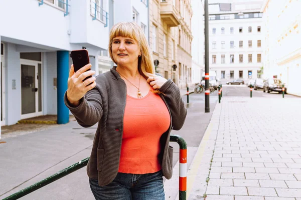 Middle-aged blonde busty woman takes selfie on smartphone camera, standing near house, against blurred background of residential modern area. Portrait of attractive middle age woman taking selfie in city