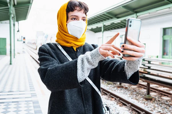 young girl in protective mask, bright yellow scarf covering hair, warm coat, stands at subway or train station, holding mobile phone. Buying tickets online, train schedules, online check-in for trips