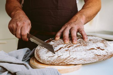 Male strong muscular hands of baker, in flour, cut loaf of craft homemade bread from rye flour. male baker at home kitchen cutting bread