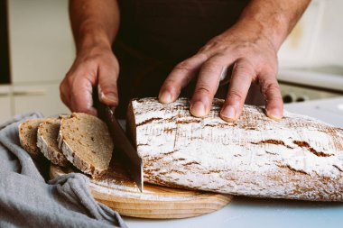 Male strong muscular hands of baker, in flour, cut loaf of craft homemade bread from rye flour. male baker at home kitchen cutting bread