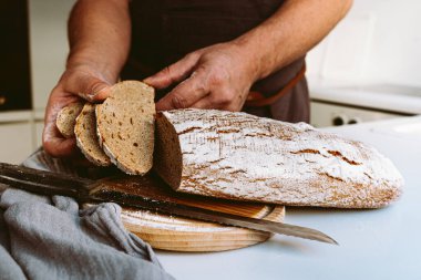 Male strong muscular hands of baker, in flour, cut loaf of craft homemade bread from rye flour. male baker at home kitchen cutting bread