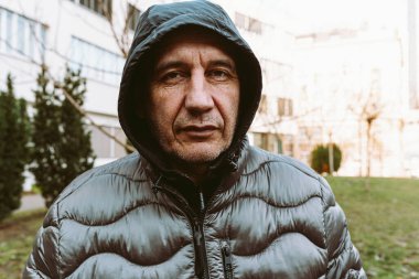 Portrait of middle-aged man in warm winter jacket with hood on blurred background of urban landscape of park. portrait of brown-eyed middle-aged man with suspicious look