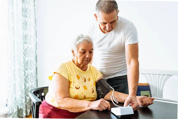 elderly woman with hypertension measures blood pressure home. middle-aged man, son stands nearby and carefully hugs mother. Sad mature overweight lady measures blood pressure with tonometer. Home care and health monitoring of elderly