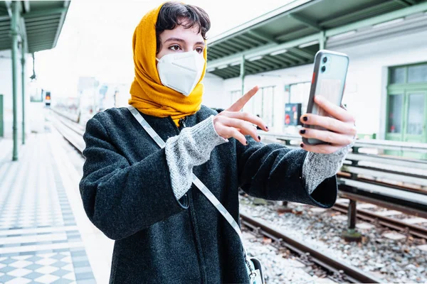 young girl in protective mask, bright yellow scarf covering hair, warm coat, stands at subway or train station, holding mobile phone. Buying tickets online, train schedules, online check-in for trips.