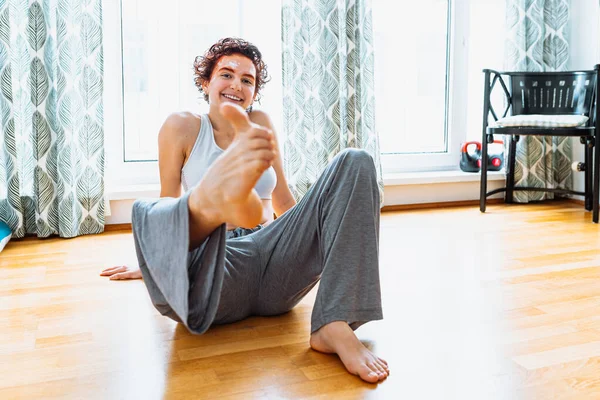 Positive yoga at home. teenager girl with red-dyed hair and eyebrows, cream mask on face, in pajamas and top, shows ok symbol with big toe, sitting on parquet floor at home.