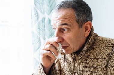 Sıcak kazaklı, burnu tek kullanımlık kağıt peçeteye sümküren, hapşıran ve öksüren orta yaşlı bir adam evde hasta. Soğuk hapşırıklardan muzdarip bir adam burnunu siliyor.