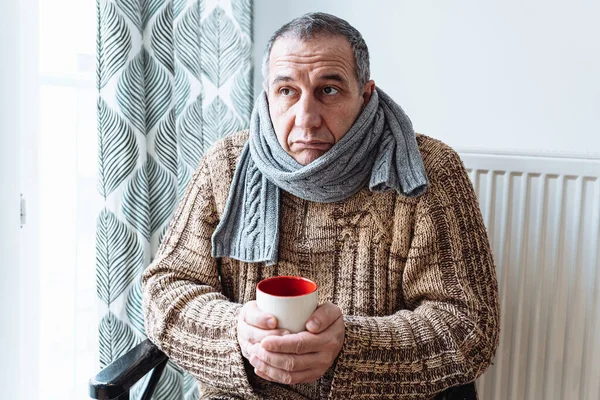 middle-aged man with cold, virus or flu, with runny nose, in warm sweater and woolen scarf, drinks warming drink from cup, an unhappy expression on face. Treatment of colds and flu with vitamin tea with lemon