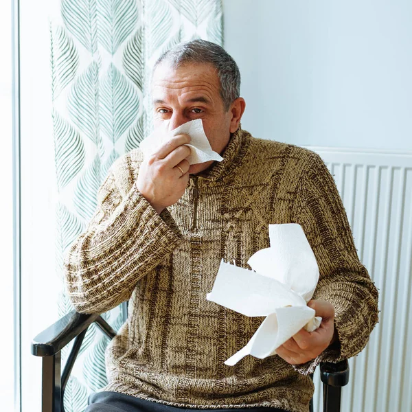 Middle-aged man in warm sweater, blows nose into disposable paper napkin, sneezes and coughs, is sick at home. Man suffering from cold sneezes and blows nose