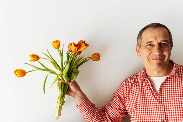 Portrait of positive smiling middle-aged man, in plaid shirt, with bouquet of tulips in hand against light wall. Man with gift bouquet positive emotion